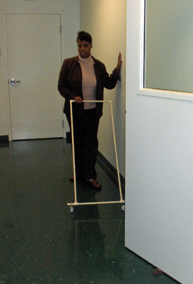 Three photos show the woman approaching the same door edge she approached in the first photos, trailing the wall with her left hand and pushing the AMD ahead of her with her right hand.  When the AMD reaches the edge of the door, it stops while the woman is still a few feet away from the door.